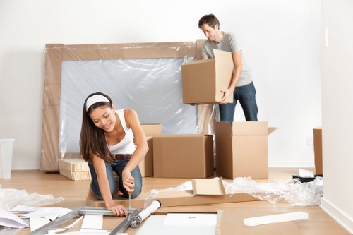 A young woman with long brown hair, wearing a white sleeveless top and a headband, is kneeling on a light wooden floor, using a utility knife to cut packaging wrap or protective material from a long cardboard box. Behind her, a young man with short dark hair, dressed in a grey t-shirt and blue jeans, is standing and carrying a medium-sized cardboard box. Several other cardboard boxes of various sizes are stacked around them, some on the floor and some on a makeshift surface. The room appears to be an interior residential space with white walls and minimal furnishings, and there is large packaging material, possibly bubble wrap, leaning against the wall in the background. The scene is well-lit, suggesting natural daylight, and the overall environment suggests a house or flat being prepared for a move in the Pimlico or central London area, consistent with services offered by Pimlico Man with Van specializing in removals in the local postcode district.
