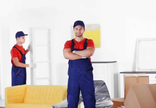 A young male removal technician from Pimlico Man with Van stands in a well-lit, modern living room during a house move, smiling and with arms crossed. He is wearing a navy blue cap, a red T-shirt, and navy blue overalls. The living room features a bright yellow upholstered sofa in the foreground, with cardboard boxes and packing materials, including a grey plastic-wrapped item, positioned nearby, indicating packing or unpacking activities. In the background, another worker is visible, wearing similar attire, moving or handling a white shelving unit against white walls with minimalistic decor. The environment appears clean and organized, with natural light illuminating the space, hinting at a residential setting in a suburban area near Pimlico, London. This scene highlights the professional approach of Pimlico Man with Van in residential removals, with focus on careful handling of household furniture and belongings, suited to the context of local house relocations within central London postal districts like SW1 or SW1V.