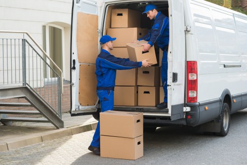 Two removal service workers dressed in blue uniforms and matching caps are loading large cardboard boxes into the back of a white commercial van parked on a residential street near a house with a staircase and metal railing. One worker is inside the van, handing a box to the other, who is standing outside on the pavement, positioned near the entrance of a property with a concrete doorstep. The boxes are plain brown with no visible labels and vary in size, with some placed on the ground nearby. The environment appears to be a suburban area, with the van positioned close to the house, suggesting a local house removal operation. The scene captures the organized activity of loading belongings for a move, reflecting professional van hire and moving services typical of areas in Pimlico or central London, emphasizing careful handling of household items during the relocation process.