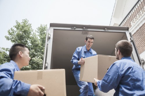 Three professional movers from Pimlico Man with Van, dressed in blue uniforms, are unloading cardboard boxes from a removals van parked outside a residential property in a suburban area, with a brick exterior and a balcony visible on the upper floor. One mover stands inside the van, handing a box to a second mover, who is standing on the ground, while the third mover to the left is carrying a box towards the vehicle. The scene takes place on a bright day, with green trees in the background and a portion of the house's white facade visible, suggesting the location is within or near Pimlico, London. The cardboard boxes vary in size, some sealed with packing tape, and are likely filled with household items being moved for a house relocation or renovation project. This imagery reflects professional house removal services, with a focus on careful handling and efficient loading, typical of removals firms operating in central London neighborhoods.