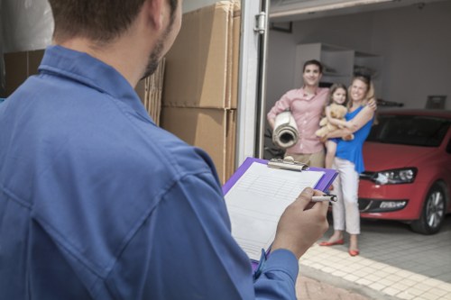 A man wearing a blue work shirt is seen from behind, holding a clipboard and pen, standing outside a residential garage. In the background, a family of three—a man, woman, and young girl—stand together near the open garage door, smiling, with the man holding a rolled-up mat, and the woman carrying a small child. A red car is parked inside the garage, and several cardboard boxes are stacked nearby. The scene appears to be part of a house removals process, with the man possibly coordinating or checking items for relocation. The setting suggests a suburban property, likely in or near Pimlico, London, consistent with local house moving services. The ambient lighting indicates daytime, and the environment appears orderly and professional, emphasizing health and safety considerations typical of removals in London neighborhoods.