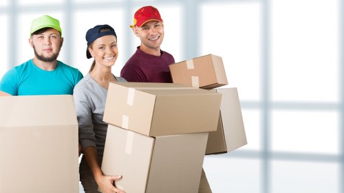 A group of three professional removalists standing indoors near a large window with natural light, dressed in casual work clothing including a green cap, a navy cap, and a red cap. They are holding and carrying multiple cardboard boxes of various sizes, some stacked and others in their hands, prepared for a house move. The boxes are plain, unmarked, and sealed with packing tape, indicating they are packed for relocation. The background shows a bright, modern interior with white walls and large windows, suggesting a residential setting, possibly a living room or hallway. The scene conveys a professional removal service environment, emphasizing careful handling and organization of household items by Pimlico Man with Van, focused on local house removals in the London area, possibly around Pimlico or nearby postcode regions. The atmosphere is orderly and focused on efficient packing and transport preparation for a house move.