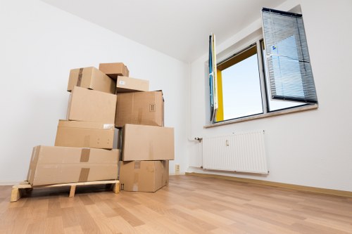 A room with light wood flooring and white walls features a stack of brown cardboard boxes of various sizes, some sealed with packing tape, arranged on a wooden pallet on the left side of the image. The boxes appear to contain household items for a house move. In the background, a window with open black blinds, partly revealing a view of a blue sky and nearby buildings, allows natural light to fill the space. Below the window, a white radiator is mounted on the wall. The room has an uncluttered, clean appearance, indicative of a recent packing process. The scene suggests a house in Pimlico or nearby in central London, with a focus on domestic removals, as provided by Pimlico Man with Van, a professional house removals service. The image is well-lit, emphasizing the textures of the cardboard, the smooth finish of the wood flooring, and the modern style of the window and radiator, representing a typical stage in a residential move within London.