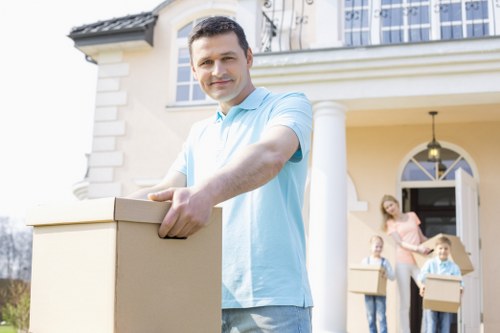 A man in a light blue polo shirt is standing outdoors in front of a large, cream-colored house with white columns, holding a cardboard moving box with both hands and smiling at the camera. In the background, two women and a young girl are seen near the entrance of the house, each carrying additional cardboard boxes, preparing for a move. The scene is set on a residential street during daytime, with a driveway and a neatly maintained garden visible. The house features a prominent front porch with a hanging lamp and large windows, reflecting a typical suburban home in London or nearby areas. The image captures the process of house removals, emphasizing careful handling of household belongings in a professional manner, consistent with the services of Pimlico Man with Van, operating within the UK.
