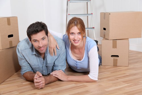 A cheerful man and woman, both with light skin, are positioned on the wooden floor of a residential interior during a house move, surrounded by several closed cardboard packing boxes of varying sizes, some with cut-out handles. The man, wearing a light blue denim shirt with dark hair, is lying face down on the floor, resting on his forearms, while the woman, with shoulder-length auburn hair and wearing a light blue and white top, is lying beside him, smiling at the camera. Behind them, there is a tall, silver aluminum ladder leaning against a white wall, and the environment appears well-lit with natural or soft ambient lighting. The scene captures a moment during a house removal in Pimlico, London, with a focus on furniture and packing materials typical of professional removals services. The floor is smooth, polished, and made of light-tone wood, contributing to a bright and organized atmosphere consistent with house relocation activities managed by Pimlico Man with Van, a company operating in central London.