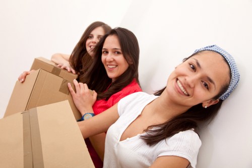 Three women involved in a house removal process, standing close together in a residential interior with white walls. The woman in the foreground, wearing a white top and a blue headscarf, is smiling and holding a cardboard moving box. Behind her, two other women are visible; one in a red top and the other in a beige top, both smiling and also supporting boxes. The boxes are of various sizes, made of brown cardboard, some with visible packing tape. The scene suggests teamwork during a household move in a typical home environment, with a clean, brightly lit space that emphasizes the physical activity of packing and moving personal belongings. The arrangement indicates the women are preparing to load or unload items, with a focus on household items such as boxes for personal belongings, possibly furniture or small appliances, in the context of house removals in the UK, specifically near Pimlico or Westminster, London.