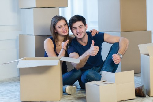 A young couple with light skin and dark hair are sitting on the floor of a well-lit room with large windows, surrounded by open cardboard boxes, some of which are partially filled with household items. The woman, wearing a sleeveless navy top, has her arm around the man's shoulder, and both are smiling at the camera. The man, dressed in a dark T-shirt and jeans, is giving a thumbs-up gesture. In the background are multiple packed moving boxes stacked neatly along the white wall, indicating an ongoing house move. The empty cardboard boxes in the foreground suggest they are in the process of packing or unpacking, typical of a home relocation in the Pimlico area. The environment appears clean and organized, reflecting a professional moving service such as Pimlico Man with Van, experienced in residential removals within central London. The scene captures a moment of satisfaction during a household move, with natural daylight illuminating the room and emphasizing the casual, yet efficient, atmosphere of the move.