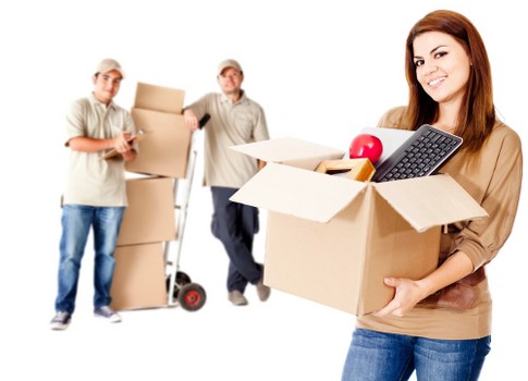 The image shows three individuals involved in a house relocation. On the left, two men are standing, each holding cardboard boxes; one man is wearing a beige cap, a light shirt, and jeans, while the other is in a similar light-colored shirt and dark trousers, with a hand truck nearby. In the foreground, a young woman with long brown hair, dressed in a beige top and jeans, is smiling and holding a cardboard box filled with household items such as a red apple, a black remote control, and a plastic container. The background is white, emphasizing the activity of moving household belongings, which include cardboard boxes and personal items, typical of house removals in Pimlico or nearby London areas. The scene captures the process of packing and preparing for a house move, reflecting a professional removals service focused on efficient and careful handling of personal possessions, with a subtle connection to recycling and sustainability aspects of moving and waste management in urban settings.
