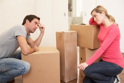 A man and a woman, both appearing to be engaged in a serious conversation, are situated in a residential interior during a house move in Pimlico. The man, wearing a light grey t-shirt and jeans, sits on the floor with a large cardboard box in front of him, leaning forward and resting his left hand on his forehead, indicating concern or frustration. The woman, dressed in a bright pink long-sleeved top and dark jeans, is seated on the floor with her right arm resting on a stack of moving boxes. She is looking at the man with a slightly concerned expression. Behind them, several filled cardboard boxes of varying sizes are stacked, some sealed with packing tape, with a few boxes placed directly on a wooden floor that shows signs of a typical home environment. The background reveals a staircase and a doorway leading to other parts of the house, illuminated by soft natural or artificial light, emphasizing the context of a home relocation process managed by Pimlico Man with Van in central London. The scene captures the moment during a house removals service, highlighting the coordination and emotions involved in a local move.