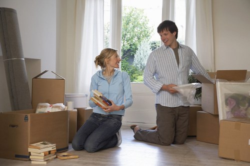 A woman and a man are inside a bright, spacious room with large windows and white curtains, preparing for a house move. The woman, seated on the wooden floor, is holding a small stack of books and smiling up at the man, who is kneeling nearby. She has blonde hair tied back and is wearing a light blue shirt and dark jeans. The man has dark hair, and is dressed in a striped shirt and beige trousers, lifting or examining a plastic-wrapped item from a partially opened cardboard box. Surrounding them, several cardboard boxes of various sizes are placed on the floor, some sealed with packing tape, others open revealing household belongings. There are also loose books stacked next to the boxes, and the scene suggests an organized moving process within a home in Pimlico or nearby in central London. The lighting is natural and bright, enhancing the clean, tidy environment typical of house removals and relocation preparations.