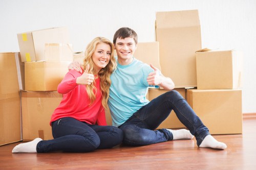 A young woman and a young man sit on a polished wooden floor inside a residential property, surrounded by numerous cardboard moving boxes of various sizes, some with packing tape and labels. The woman has long blonde hair and is wearing a red top and dark blue jeans, while the man has short dark hair and is dressed in a light blue t-shirt and dark jeans. Both are smiling and giving thumbs-up gestures, indicating a successful move. In the background, there is a plain white wall, and the scene suggests an organized household move typical of London or surrounding areas, with the focus on the furniture and packed belongings ready for transport. The natural lighting highlights the textures of the cardboard boxes, the wooden floor, and the casual clothing, conveying a positive and professional atmosphere associated with house removal services by Pimlico Man with Van.