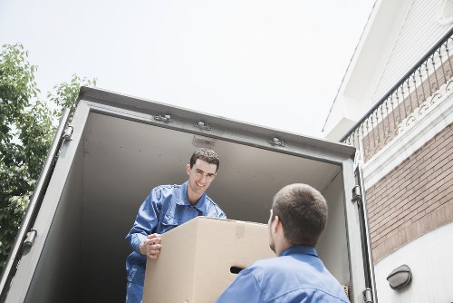 Two professional movers from Pimlico Man with Van, dressed in blue uniforms, are loading a large cardboard box into the back of a moving van parked outside a residential property in Pimlico. The interior of the van is visible, showing a spacious cargo area suitable for household belongings. One mover, with a focused expression, is handing the box to the other who has his back to the camera. The box appears to be sturdy and plain, made of brown corrugated cardboard, suitable for house removals. The background includes part of a brick house with a white exterior wall and a balcony railing on the right, along with a glimpse of greenery from trees nearby. The scene is illuminated by natural daylight, suggesting daytime conditions. This image captures the typical process of house removals in London, highlighting careful handling and loading of household items, consistent with services offered by Pimlico Man with Van in central London neighborhoods such as Pimlico and surrounding postcode areas.