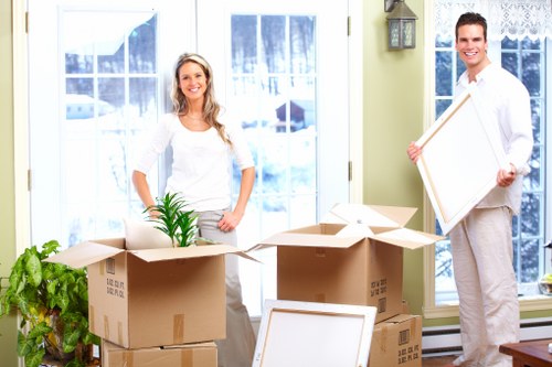A smiling woman with long blonde hair, wearing a white top and light grey trousers, stands in a bright, well-lit room with large windows and snowy outdoor scenery visible through the glass. She is positioned behind a collection of cardboard moving boxes, some open and filled with household items, including a small green potted plant. To the right, a man with short dark hair, dressed in a white shirt and light trousers, holds a large picture frame, also smiling. The room features a light wall with a small decorative lantern and a leafy houseplant near the boxes. The scene captures an indoor residential space prepared for a house move, with items arranged on the floor ready for transportation, and the natural daylight enhancing the professional yet casual atmosphere typical of house removals in the UK, such as in Pimlico or nearby areas.