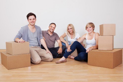 Four individuals are seated on a light wooden floor inside a residential space, surrounded by various cardboard moving boxes in different sizes, some stacked on their sides and others placed flat. The group consists of two men and two women, all casually dressed in neutral and light-colored clothing. The man on the far left wears a light blue shirt and beige trousers, smiling with one hand resting on a box, while the man next to him is in a grey t-shirt and jeans, also smiling and placed between the women. The woman on the right wears a white sleeveless top and dark jeans, sitting with her legs crossed and a relaxed expression, and the woman in the middle has shoulder-length blonde hair, a white top, and dark trousers, smiling at the camera. In the background is a plain white wall, giving the impression they are in a home or flat in Pimlico, London, preparing for a house move. The scene suggests a team or family involved in house removals or packing, supported by the presence of multiple boxes and a tidy, organized environment, representative of removals services offered by Pimlico Man with Van in the area.