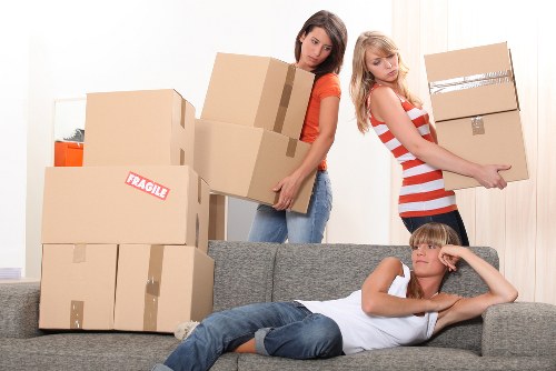 In a residential interior, three individuals are involved in packing and moving household belongings. Two young women, one with long brown hair wearing a red top and jeans, and the other with blonde hair in a red and white striped dress, are carefully carrying cardboard boxes, some labeled with tape or markings, towards a door or hallway, indicating preparations for a house relocation. Several stacked cardboard boxes of varying sizes, some sealed with packing tape and others open or partially packed, are seen on a grey fabric sofa in the foreground. A young boy with blonde hair, lying on the sofa, rests his head on his hand and appears to be observing the activity with a casual expression. The room is well-lit with natural light filtering through curtains, and the background features a patterned wall and a hallway entrance, suggesting a typical UK home, potentially in Pimlico or nearby London areas. The scene overall depicts the process of house removals, with attention to household items, furniture, and packing materials typical of a professional removals service in the local area.