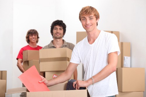 In a brightly lit interior space, three movers from Pimlico Man with Van are positioned amidst an array of cardboard boxes, likely in a residential property in the Pimlico area of central London. The young man in the foreground, wearing a white t-shirt, is smiling and holding a red envelope or document while standing next to partially packed boxes. Behind him, two other movers—a woman dressed in a red top and a man with dark curly hair and a beige shirt—are also smiling and carrying stacked medium-sized cardboard boxes with printed labels. The boxes are positioned on a table or workspace, with some stacked in the background, indicating an ongoing or recent packing process. The environment appears to be a clean, organized house interior or hallway, suitable for a house removal service. The scene captures a professional, cooperative moment during residential furniture and personal item relocation, reflecting the reliability and efficiency of Pimlico Man with Van's removals services in London's central postcode areas, such as SW1 or SW1V, consistent with the company's location and service focus.