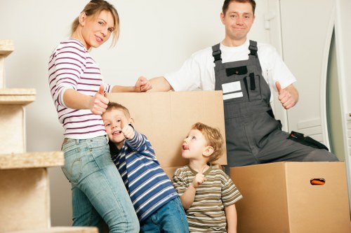 A family preparing for a house move indoors, with a woman, children, and a man in work overalls standing in front of packed cardboard boxes. The woman is wearing a striped long-sleeved top and jeans, while the two young children, a boy and a girl, are dressed in casual striped T-shirts, smiling and giving thumbs-up signs. The man, clad in grey work overalls over a white T-shirt, is smiling and standing next to the boxes, which are positioned on a staircase and on the floor of a residential interior. The scene is well-lit, with the background showing a white wall and a doorframe, indicating a typical house setting in Pimlico or nearby London, preparing for a local move or house removal by Pimlico Man with Van, a professional removals company specializing in local residential relocations. The image captures the preparations involved in a household move, emphasizing family involvement and organized packing within a typical home environment.