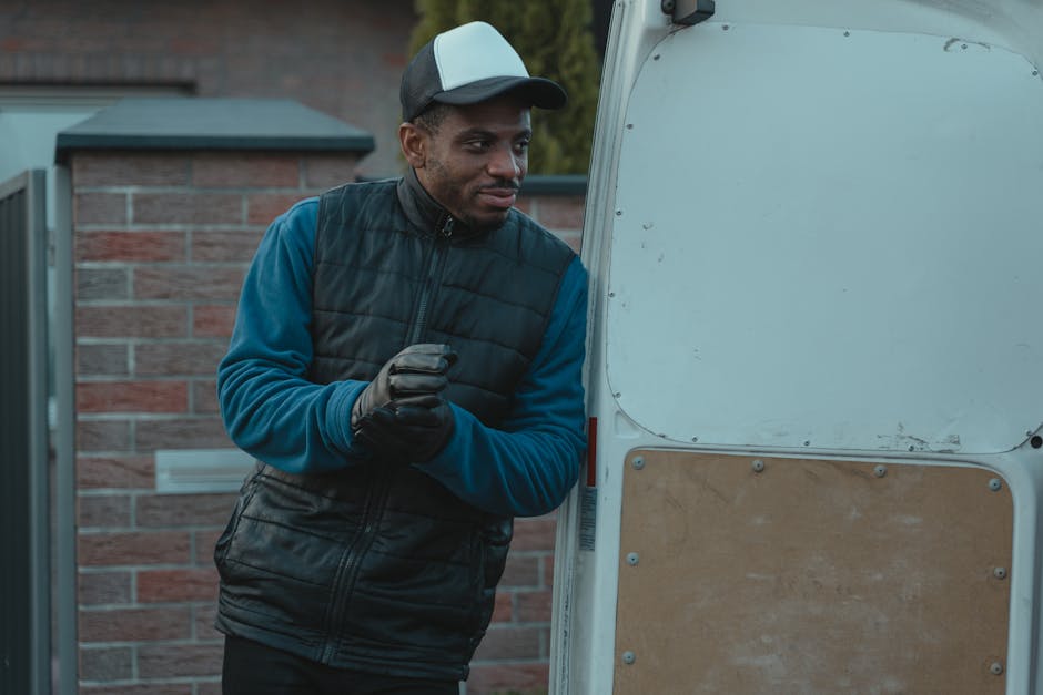 A man wearing a black and gray cap, a dark padded vest over a blue long-sleeve shirt, and black gloves stands outside next to the rear of a white moving van. The van's open door reveals part of its in