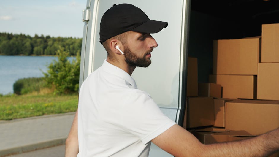 A man wearing a black and gray cap, a dark padded vest over a blue long-sleeve shirt, and black gloves stands outside next to the rear of a white moving van. The van's open door reveals part of its in