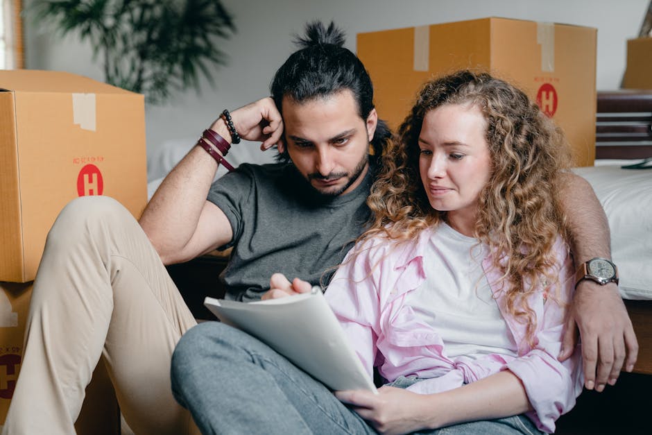 A woman with curly hair tied back in a bun is inside a room, leaning over a large cardboard box on a table. She is writing a packing or inventory list with a black marker on the surface of the box. Th